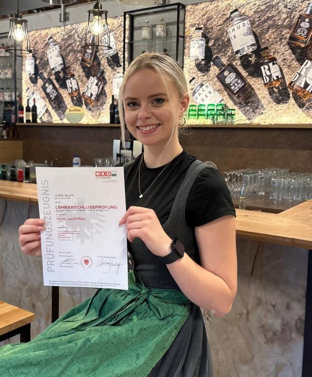 Junge Frau im Café hält eine Prüfungsurkunde zur bestandenen Lehrabschlussprüfung als Hotelkauffrau.