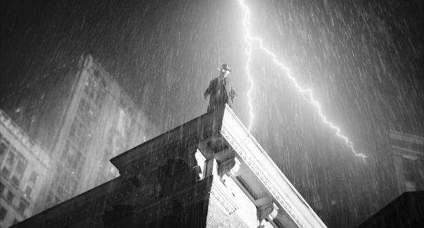 Eine in Trenchcoat und Hut gekleidete Gestalt steht bei strömendem Regen und einem Blitzschlag auf einem Gebäudedach.