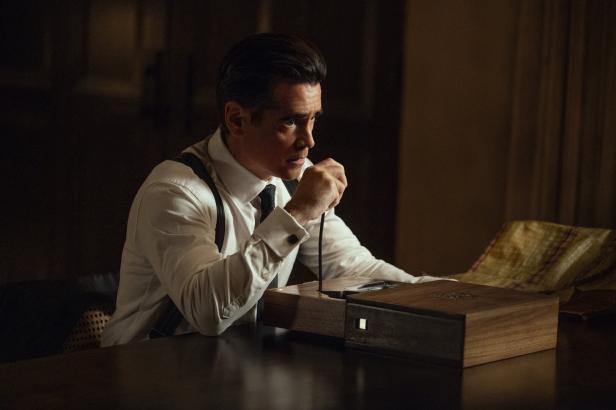 Colin Farrell sitzt in einem weißen Hemd mit Hosenträgern an einem Tisch und hält ein Kabel, das mit einem hölzernen Gerät verbunden ist.