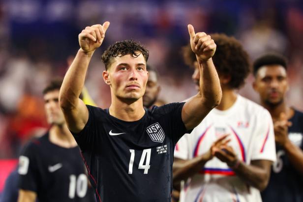 Ein US-Fußballspieler mit der Trikotnummer 14 hebt beide Daumen in die Höhe, während er vor seinen Teamkollegen im Stadion steht.
