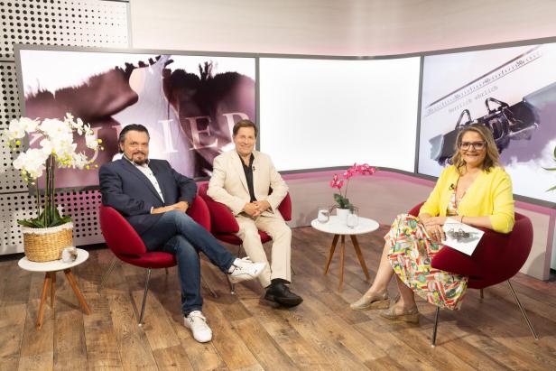 Zwei Männer und eine Frau sitzen in einem Fernsehstudio in roten Sesseln vor Bildschirmen und unterhalten sich.