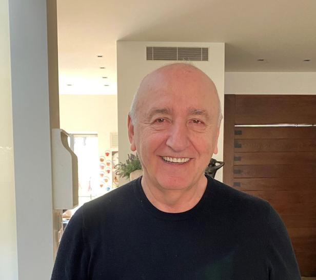 Lachender älterer Mann mit Glatze und schwarzem Pullover in einem Innenraum.