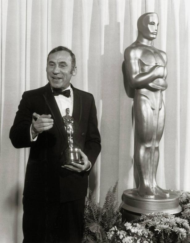 Mel Brooks im Smoking hält einen Oscar in der Hand, während er neben einer großen Oscar-Statue steht.