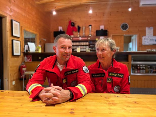 Zwei Personen in roten Rettungsjacken sitzen an einer Holztheke in einer Hütte und lächeln sich an.