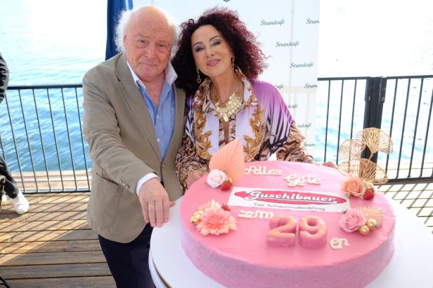 Ein älteres Paar posiert am Meer vor einer großen rosa Geburtstagstorte mit der Aufschrift „Alles Gute zum 29.“.