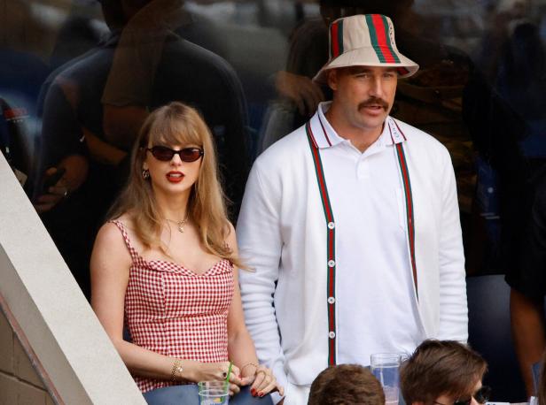 Taylor Swift und Travis Kelce sitzen nebeneinander auf einer Tribüne, wobei sie ein rot-weiß kariertes Kleid und er einen Gucci-Hut trägt.