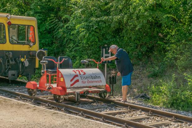 Mann schiebt eine Draisine neben einem gelben Zug auf den Schienen.