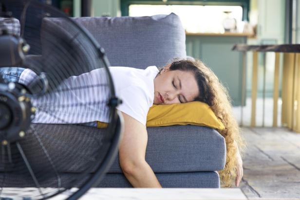 Eine Frau schläft erschöpft auf einer Couch, ihr Kopf liegt auf einem gelben Kissen, davor ein laufender Ventilator.