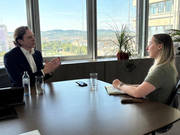 Ein Mann und eine Frau sitzen sich an einem Konferenztisch vor einem großen Fenster mit Blick auf eine Stadt gegenüber.