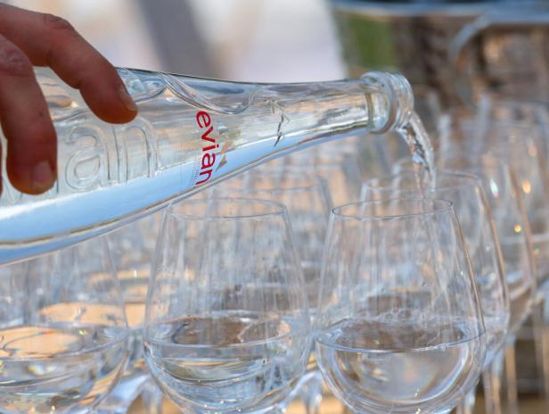 Eine Hand gießt Evian-Mineralwasser aus einer Glasflasche in mehrere bereitstehende Weingläser.