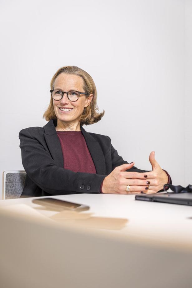 Eine lächelnde Frau mit Brille sitzt am Tisch und gestikuliert im Gespräch.