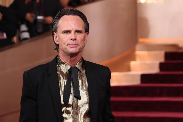 Walton Goggins trägt ein goldenes Hemd mit schwarzer Krawatte und ein schwarzes Sakko vor einer Treppe mit rotem Teppich.