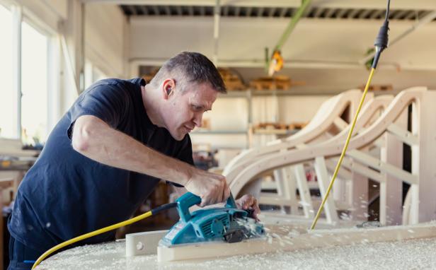 Ein Tischler bearbeitet in einer Werkstatt ein Holzstück mit einem elektrischen Handhobel.