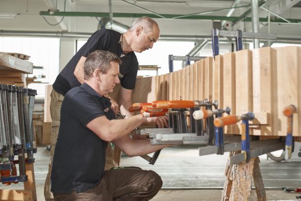 Zwei Männer in einer Werkstatt spannen Holzteile mit vielen Schraubzwingen zusammen.
