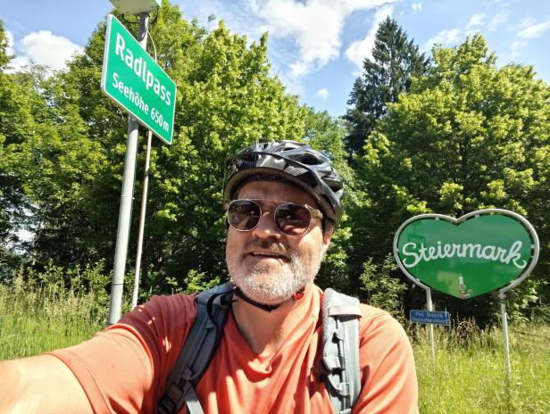 Ein Radfahrer mit Helm und Sonnenbrille posiert vor einem Schild am Radlpass in der Steiermark.