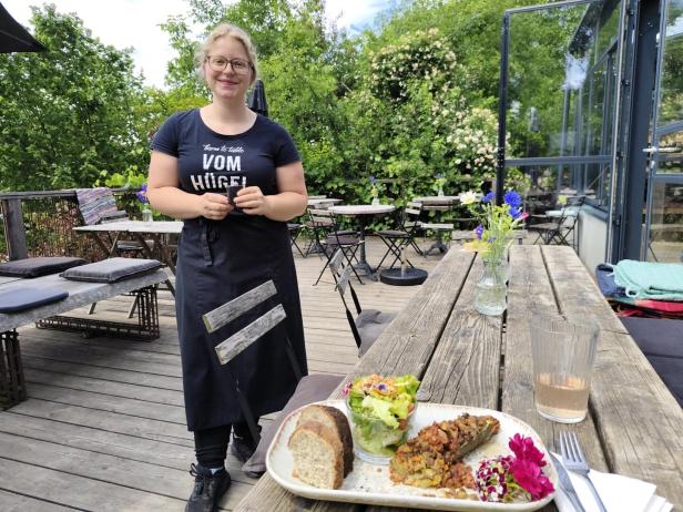 Kellnerin steht auf einer grünen Terrasse vor Tischen; im Vordergrund liegt ein vegetarischer Teller mit Brot und Salat.