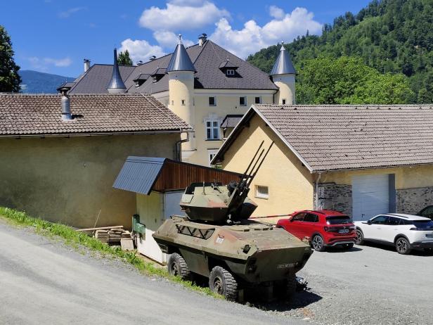 Ein gepanzerter Fahrzeugwagen steht vor Häusern, dahinter ein schlossartiges Gebäude und bewaldete Berge.