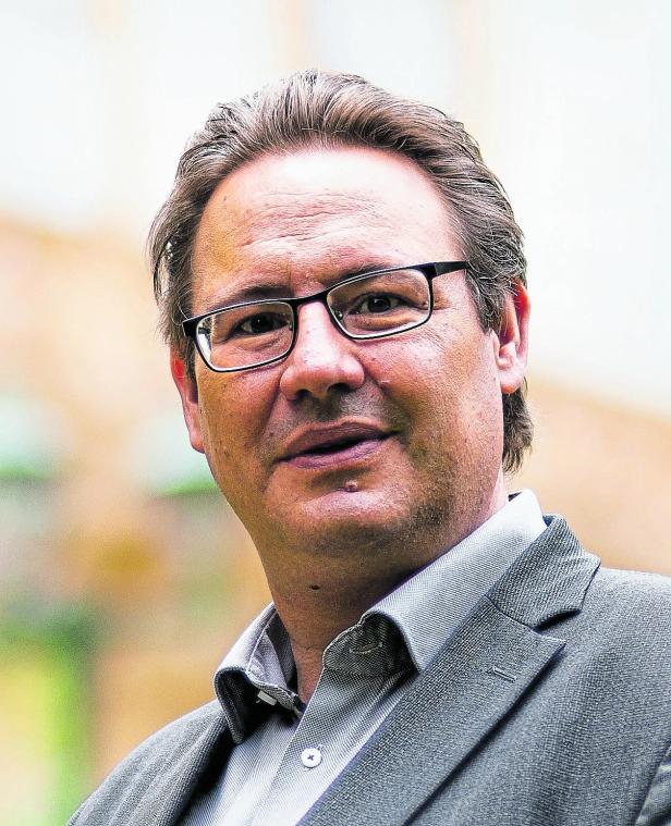 Nahaufnahme eines Mannes mit Brille im grauen Anzug vor unscharfem Hintergrund.