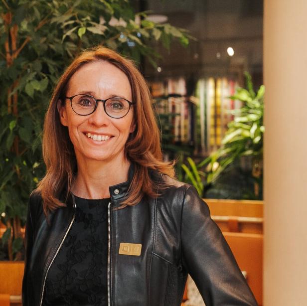 Lächelnde Frau mit Brille und schwarzer Lederjacke in einem begrünten Innenraum.