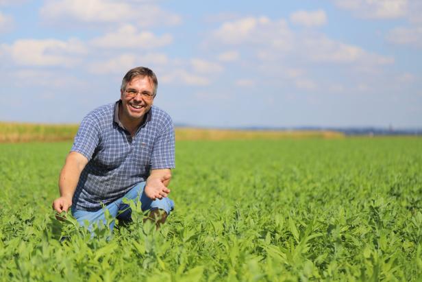 Ein lächelnder Mann kniet in einem grünen Feld unter blauem Himmel mit Wolken.