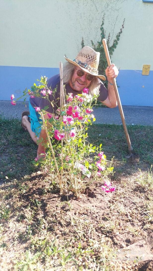 Ein Mann mit Strohhut kniet im Garten und arbeitet mit einer Hacke an einem blühenden Strauch.