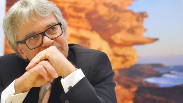 Ein Mann mit Brille und Anzug vor einem unscharfen Hintergrund mit Felsen und Meer.