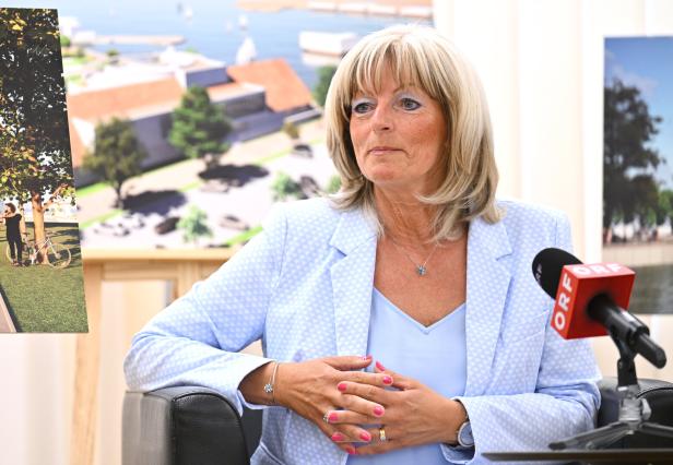 Eine blonde Frau in einem hellblauen Blazer sitzt vor einem ORF-Mikrofon und präsentiert architektonische Entwürfe.