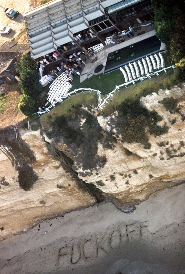 Luftaufnahme eines Malibu-Anwesens an der Klippe mit aufgebauter Hochzeitsdekoration und dem in den Sand geschriebenen Wort „FUCK OFF“.