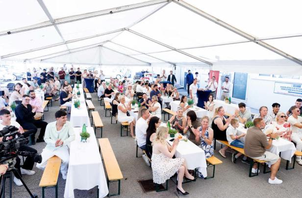 Menschen sitzen an langen Tischen unter einem großen weißen Festzelt und klatschen.