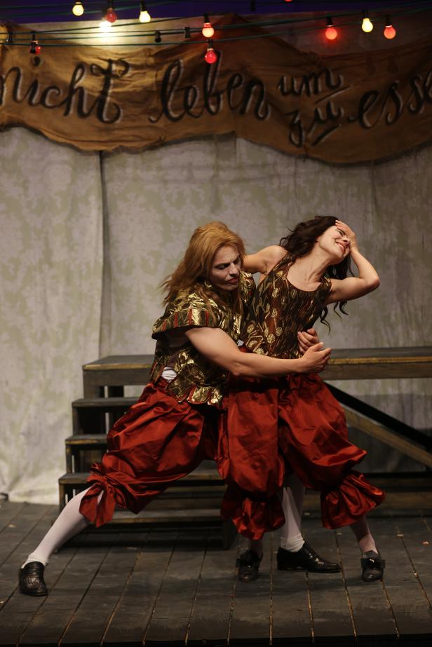 Zwei Darstellerinnen in roten Kostümen umarmen sich dramatisch auf einer Theaterbühne mit Lichterkette.