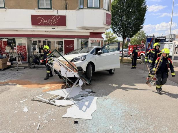 Auto prallt gegen zerstörtes Ladenschild vor einem Geschäft; Feuerwehrleute sichern die Unfallstelle.