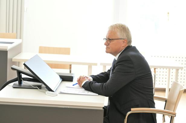 Älterer Mann im Anzug sitzt an einem Tisch und hört aufmerksam vor einem Bildschirm zu.