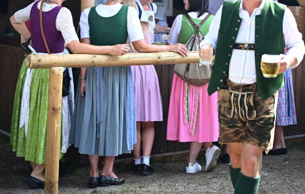 Menschen in traditioneller Tracht stehen an einem Holzzaun, während ein Mann mit Bierkrug im Vordergrund vorbeiläuft.