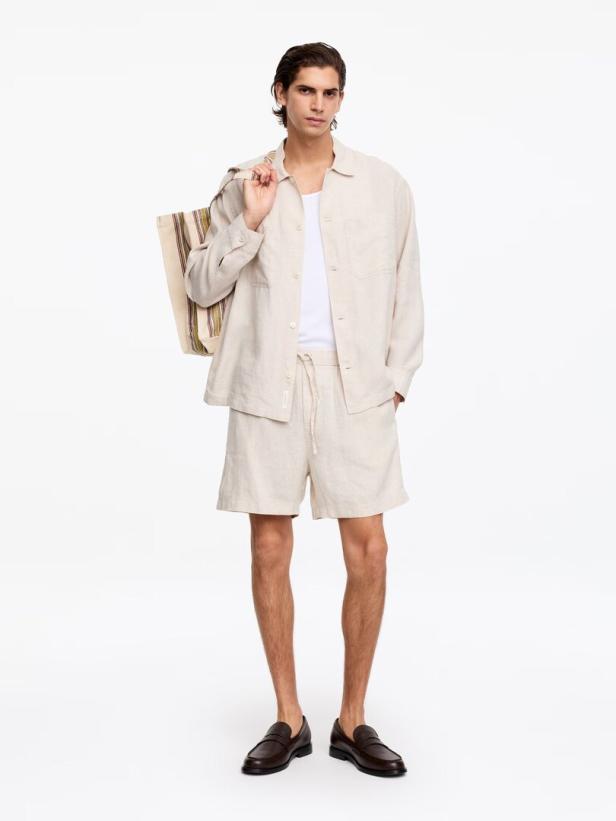 Ein Mann trägt ein beiges Leinenhemd, ein weißes Unterhemd, passende Shorts und eine gestreifte Tasche über der Schulter vor weißem Hintergrund.