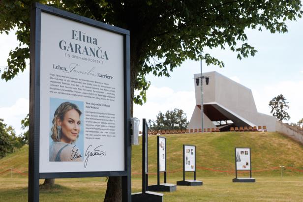 Ausstellungstafeln zu Elīna Garanča stehen auf einer Wiese vor einem modernen Gebäude unter Bäumen.