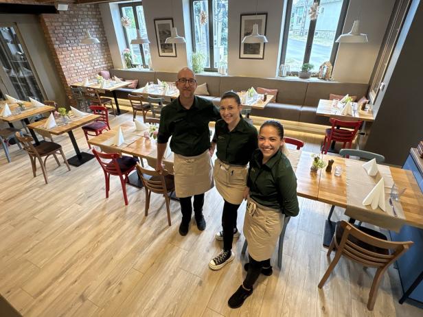 Drei Restaurantmitarbeiter stehen lächelnd in einem hellen, modern eingerichteten Gastraum.