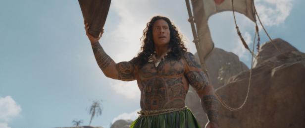 Dwayne Johnson als tätowierter Halbgott Maui hält einen Gegenstand in die Höhe, während er auf einem Boot vor einer felsigen Küste steht.