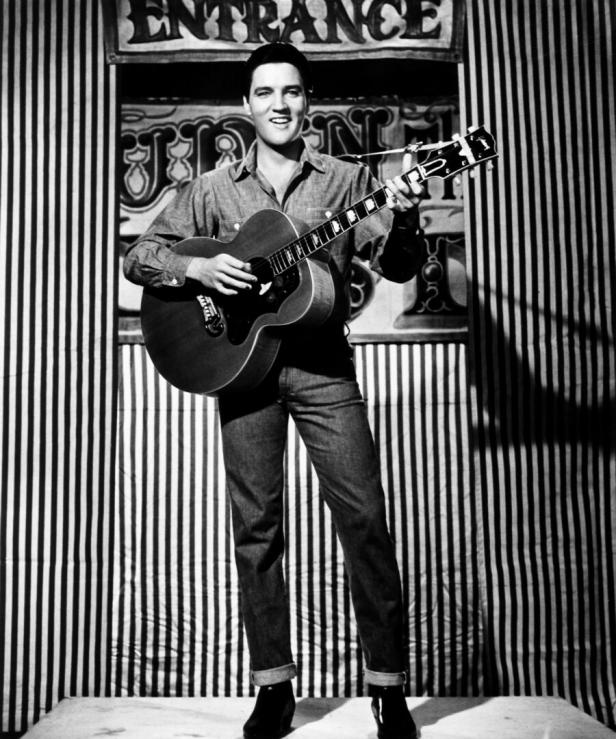 Elvis Presley steht mit Gitarre vor gestreiftem Bühnenhintergrund.