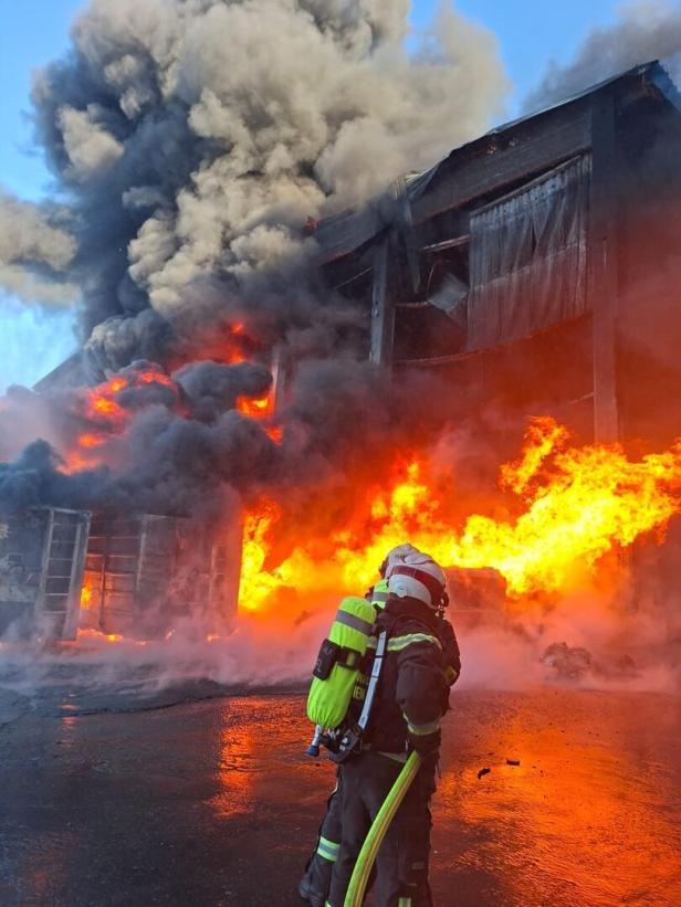 Feuerwehrmann vor einem brennenden Gebäude mit dichtem Rauch und meterhohen Flammen.