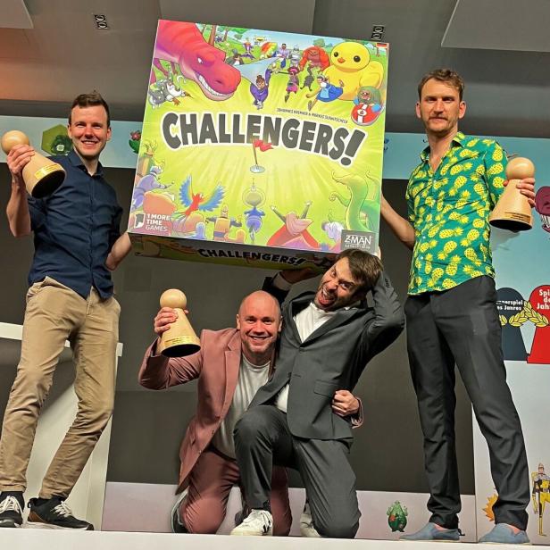 Vier Männer posieren mit einer großen Challengers!-Spielebox und Holzpokalen vor einer Messewand.