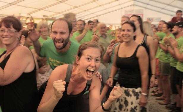 Eine jubelnde Menschenmenge in einem Festzelt, wobei eine Frau im Vordergrund begeistert die Fäuste ballt und ein Mann im grünen T-Shirt laut mitfeiert.