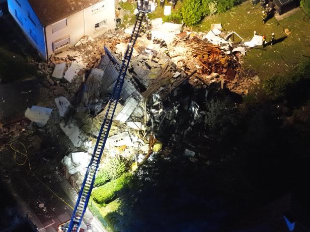 Eingestürztes Haus mit Trümmern, Feuerwehrleuten und einer ausgefahrenen Drehleiter bei Nacht.