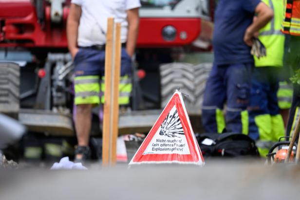 Ein Warnschild mit der Aufschrift „Explosionsgefahr“ steht vor Bauarbeitern und einem Baufahrzeug.