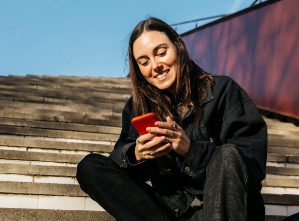 Junge Frau sitzt auf Treppen und lächelt auf ihr rotes Smartphone.