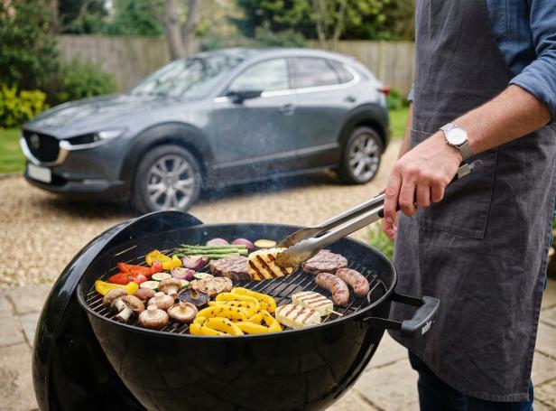 Person grillt Gemüse, Fleisch und Würstchen auf einem Kugelgrill im Garten, im Hintergrund steht ein Auto.