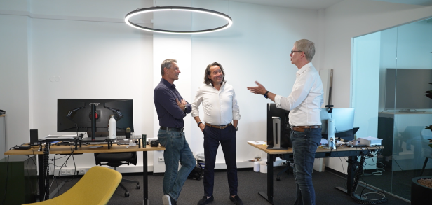 Drei Männer unterhalten sich in einem modernen Büro zwischen Schreibtischen und Monitoren.