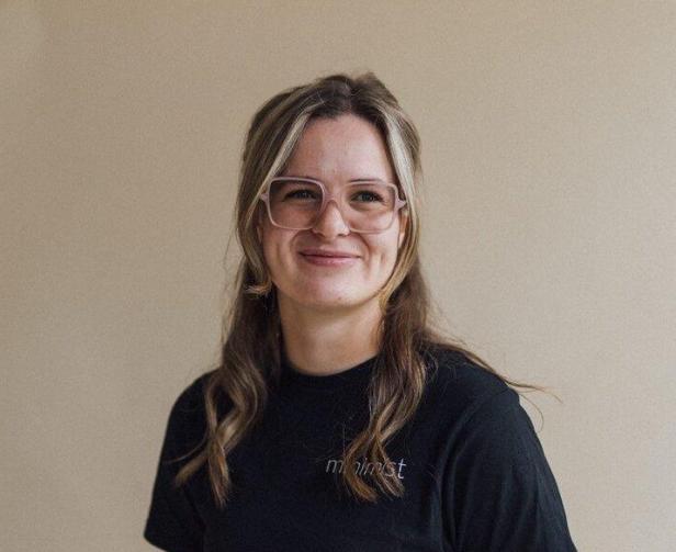 Eine lächelnde Frau mit Brille und dunklem T-Shirt steht vor einer hellen Wand.