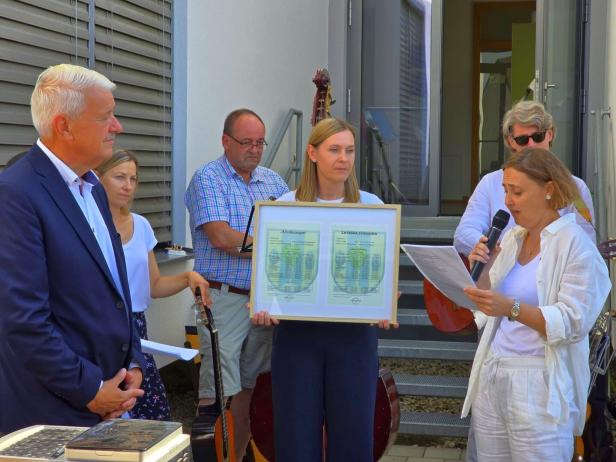 Eine Frau liest vor einer Gruppe draußen aus einem Papier vor, während zwei Frauen ein gerahmtes Dokument halten.