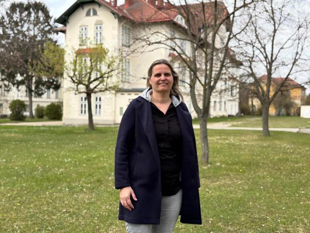 Frau steht im Park vor einem hellen Haus mit roten Dächern und kahlen Bäumen.