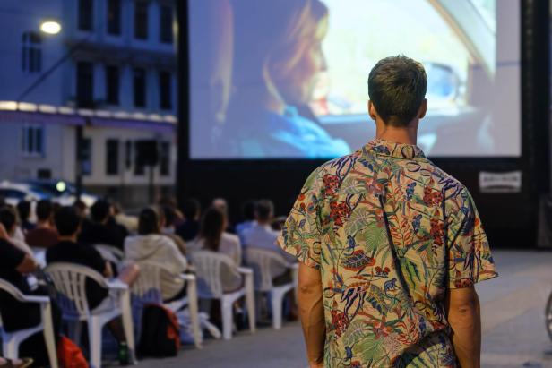 Mann im bunten Hemd schaut auf eine Filmvorführung im Freien mit sitzendem Publikum.